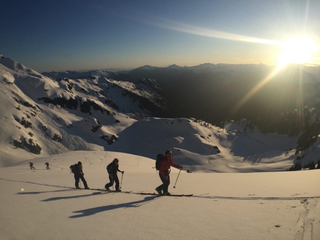 A ski-tour group makes its way across the slope as the sun sets behind the mountain.